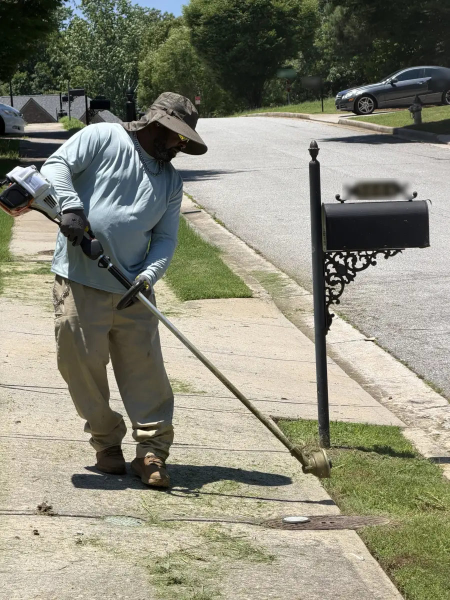 Worker in a wide hat using a string trimmer to edge grass along a sidewalk by a mailbox