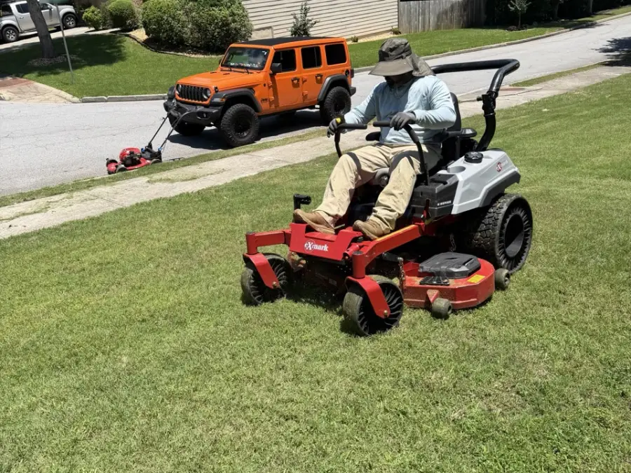 Rider on a red zero-turn mower crossing a green front lawn near the street